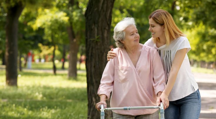 Pflegekraft und Seniorin gehen im sommerlichen Park spazieren